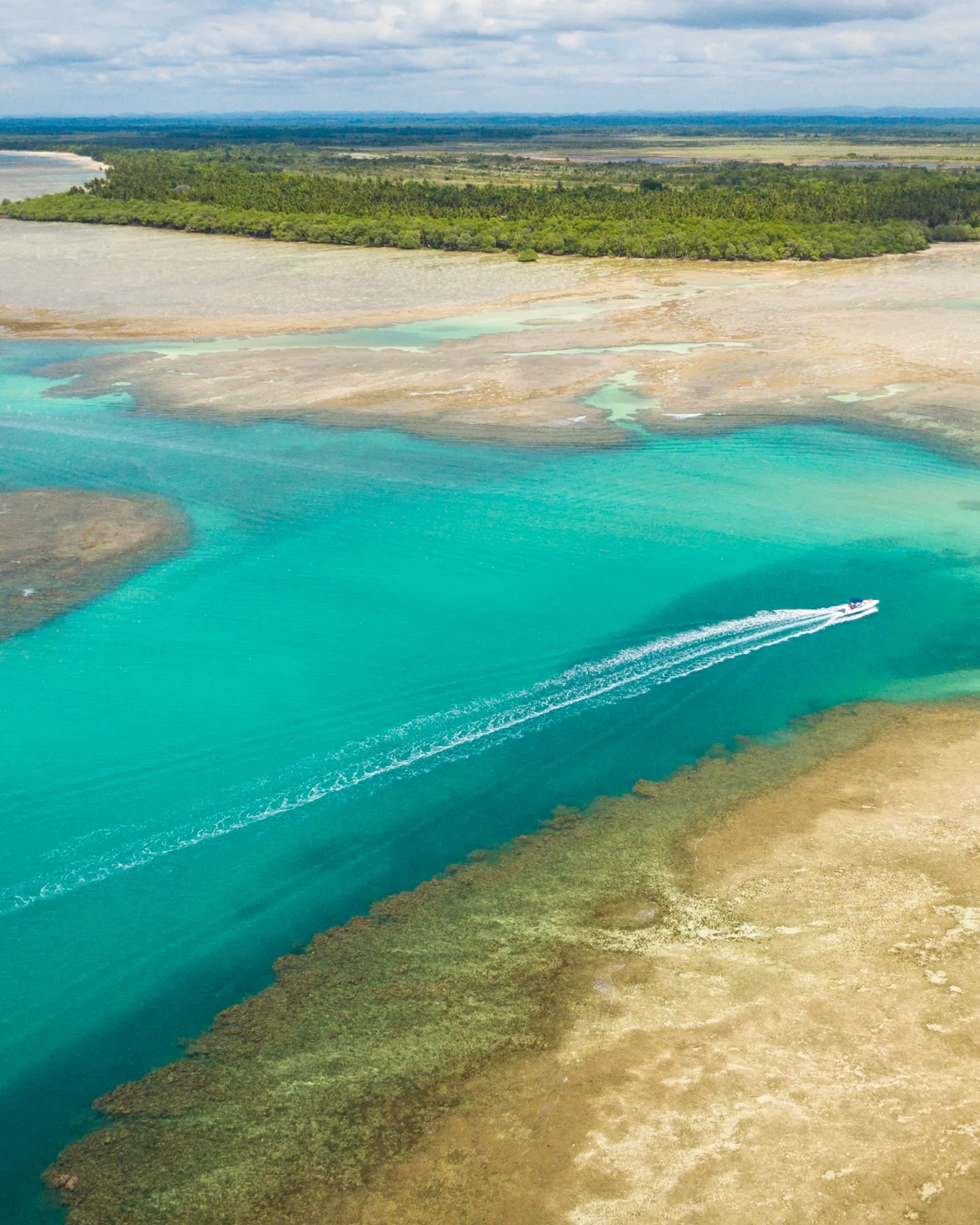Vista aérea do canal turquesa de Moreré, na Ilha de Boipeba, na Bahia, com bancos de areia, recifes e uma lancha deixando um rastro de espuma.