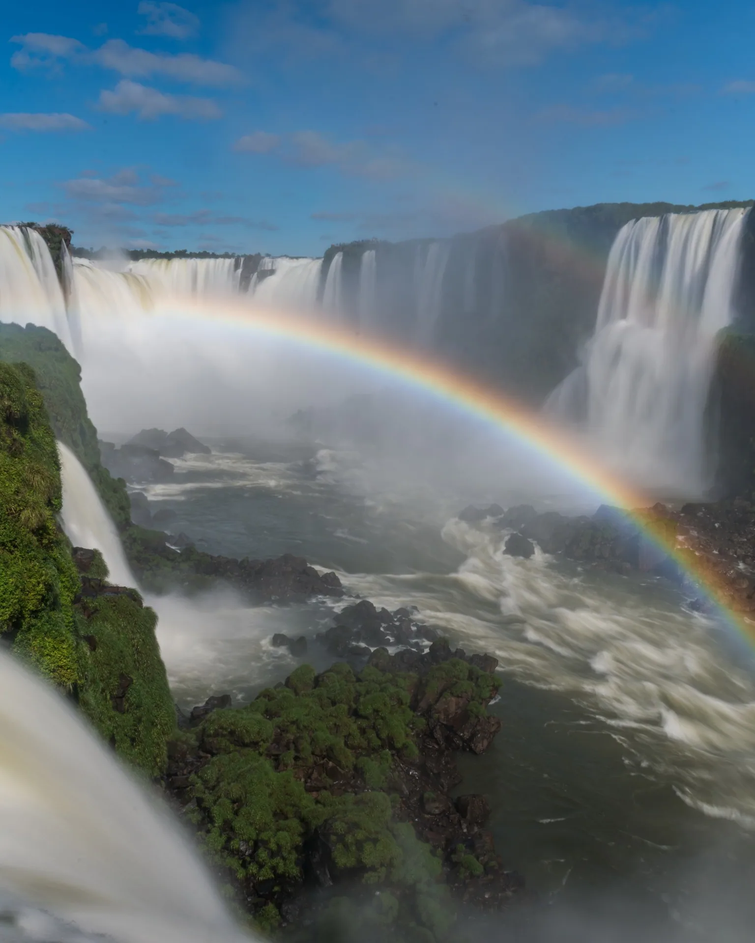 Quedas das Cataratas do Iguaçu cercadas de mata, com um arco-íris sobre a névoa.