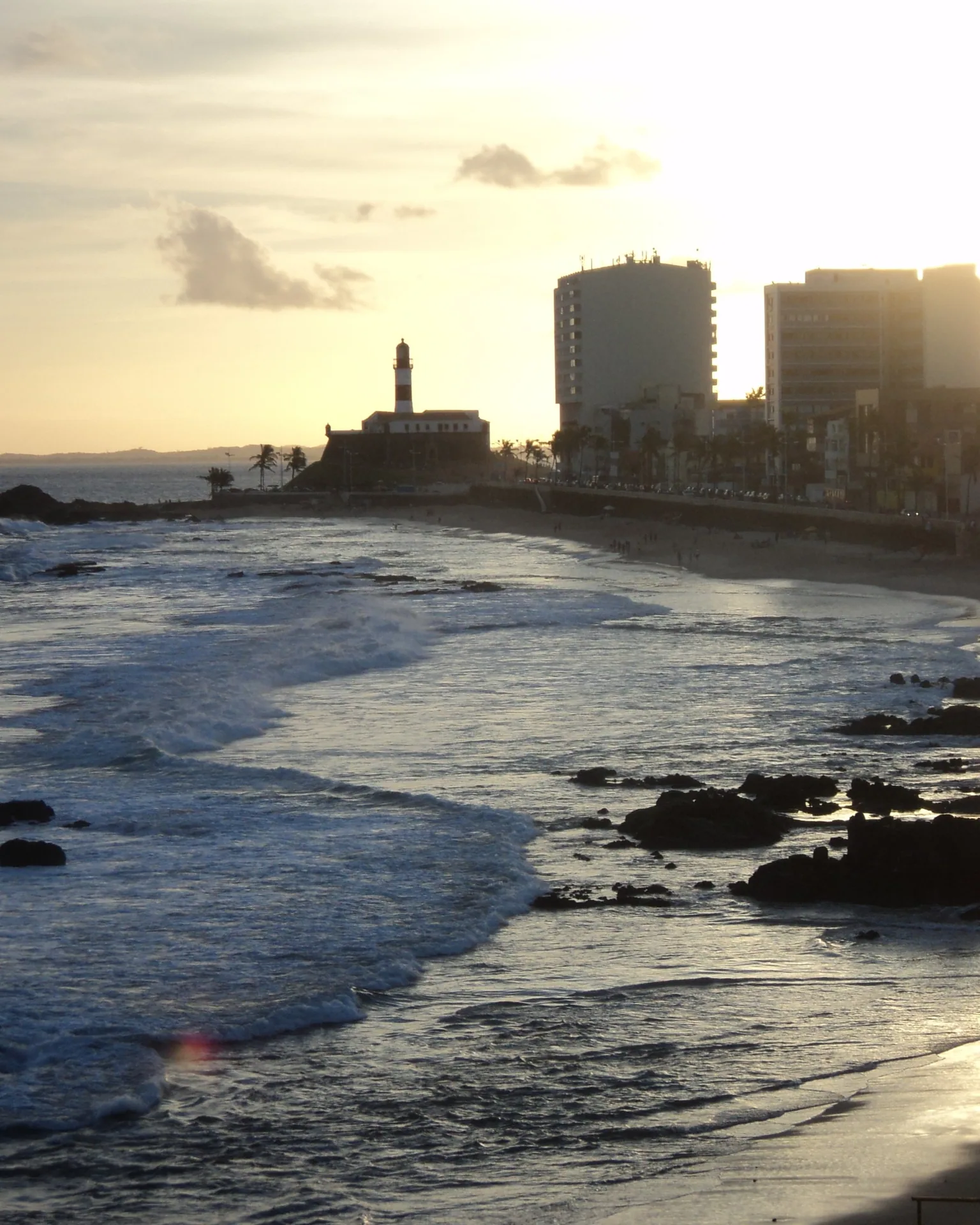 Pôr do sol na praia do Farol da Barra, em Salvador, com ondas na areia e o farol ao fundo.