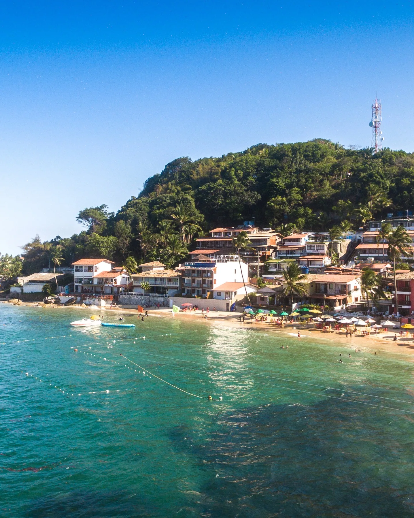 Primeira Praia de Morro de São Paulo, na Bahia: casas e pousadas no morro verde, guarda-sóis na areia e mar turquesa.