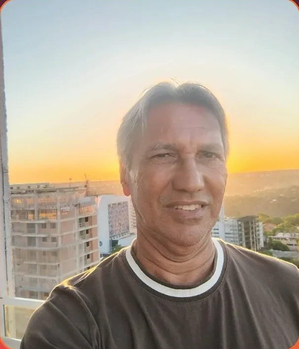 Retrato de Solivam Tagarro, de cabelo grisalho e camiseta marrom, ao pôr do sol com prédios ao fundo.